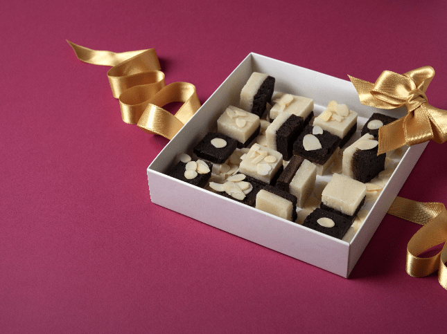 Scatola di dolcetti bicolore con scaglie di mandorle e nastro dorato su sfondo bordeaux